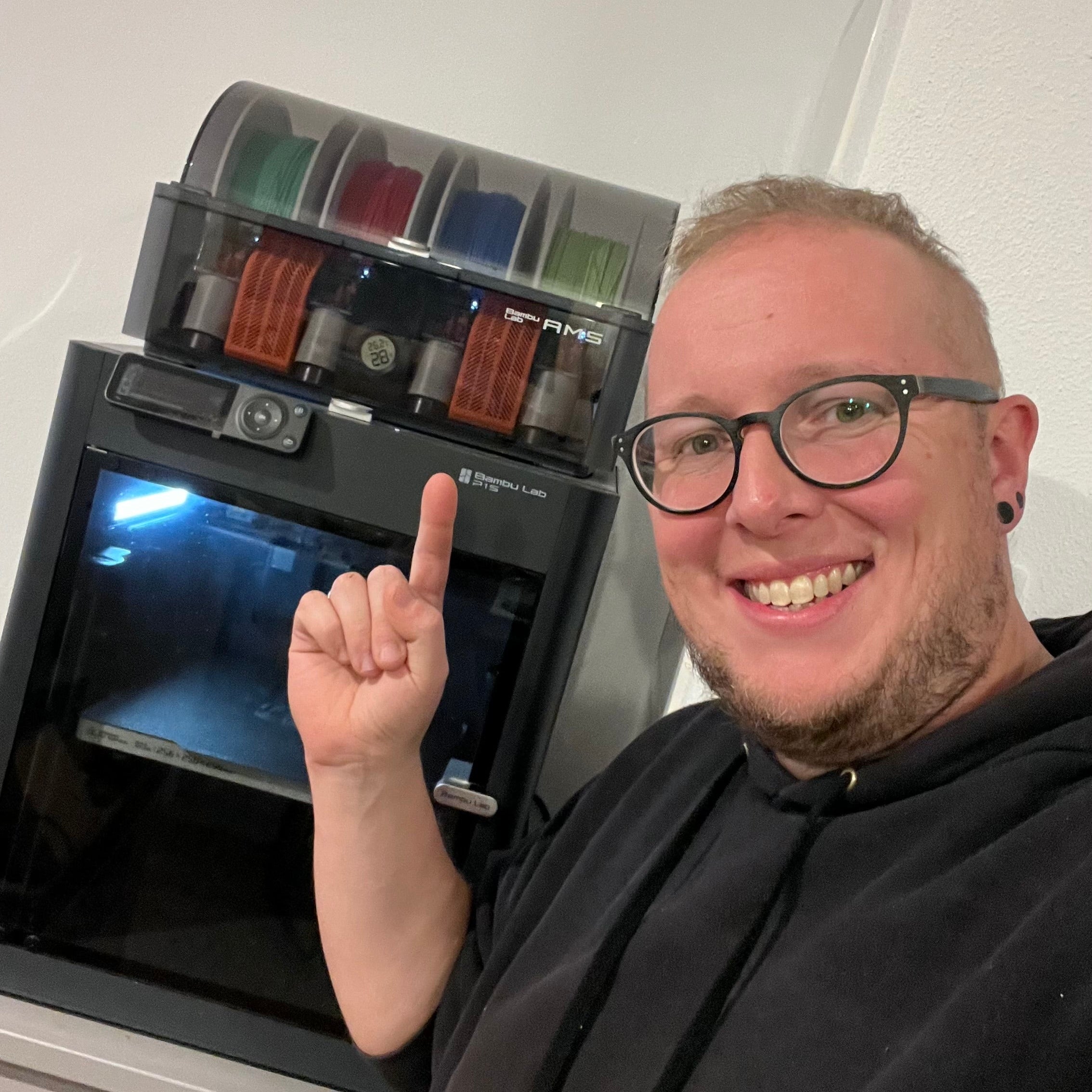 Martin Sämeier steht vor seinem 3D Drucker und zeigt darauf. Zu sehen ist der Bambu Lab P1S mit dem AMS darauf. Im AMS sind die Farben Grasgrün, Rot, Blau und Apfelgrün