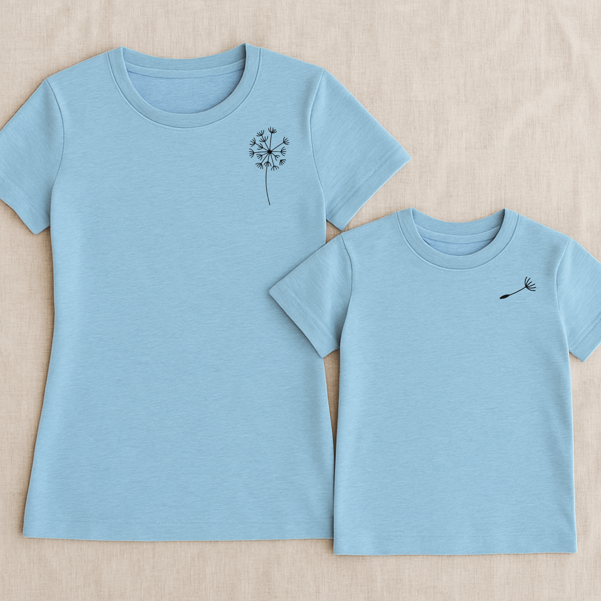 Mama-Kind-Shirt-Set aus Bio-Baumwolle mit Pusteblumen-Design: Auf dem taillierten Mama-Shirt fehlt ein Schirmchen, das auf dem Kind-Shirt einzeln erscheint. Symbol für Liebe, Verbindung und Loslassen. Die Shirts liegen auf natürlichem Hintergrund, modern und nachhaltig inszeniert. Ideal als Muttertagsgeschenk, zur Geburt oder für emotionale Familienmomente. Hier Set in hellblau.