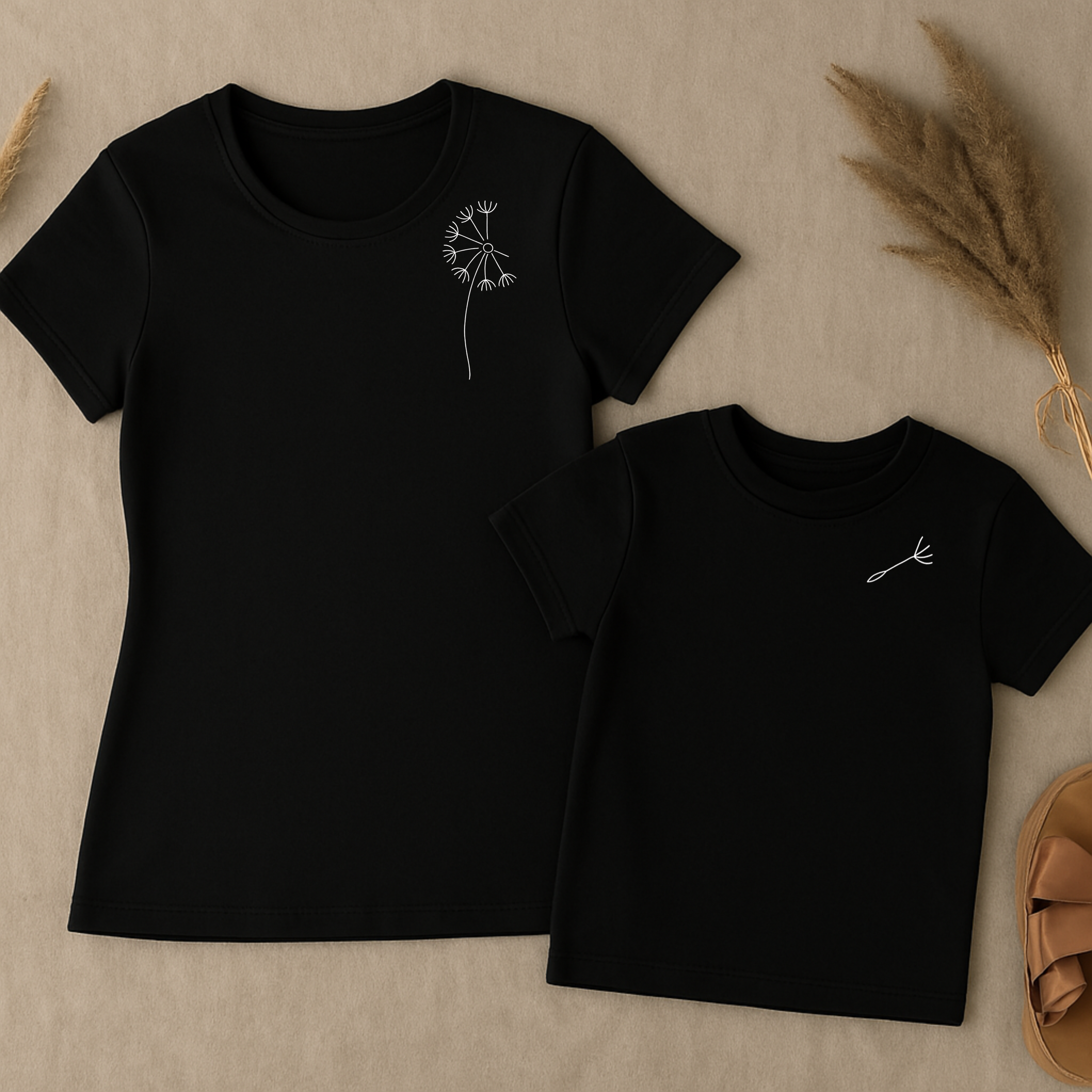 Mama-Kind-Shirt-Set aus Bio-Baumwolle mit Pusteblumen-Design: Auf dem taillierten Mama-Shirt fehlt ein Schirmchen, das auf dem Kind-Shirt einzeln erscheint. Symbol für Liebe, Verbindung und Loslassen. Die Shirts liegen auf natürlichem Hintergrund, modern und nachhaltig inszeniert. Ideal als Muttertagsgeschenk, zur Geburt oder für emotionale Familienmomente. Hier Set in schwarz